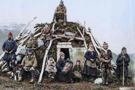 Een groep mensen in traditionele kleding poseert voor een met aarde bedekte woning. Sommigen zitten, anderen staan, met kinderen en een hond tussen hen in. Op de achtergrond een met sneeuw bedekt landschap.